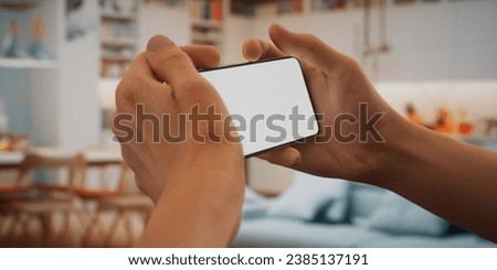CU Caucasian man using his phone at home. Daytime shot, furnished house interior in the background. Blank screen, smartphone mockup