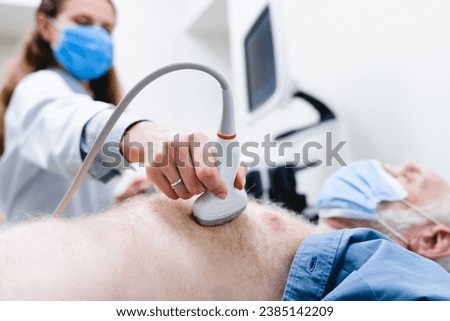 Focused photo of ultrasound scanner checking mature patient`s chest by female doctor in mask in the background. Cardiovascular illness disease prevention. Heartbeat Royalty-Free Stock Photo #2385142209