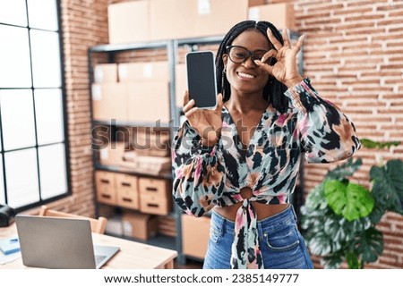 African woman with braids working at small business ecommerce showing smartphone screen smiling happy doing ok sign with hand on eye looking through fingers 