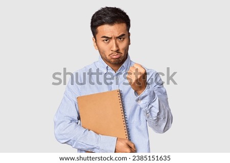 Chinese student with a notebook in studio showing fist to camera, aggressive facial expression.