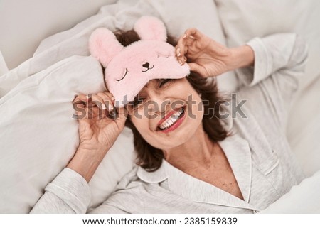 Middle age woman wearing funny sleep mask lying on bed at bedroom