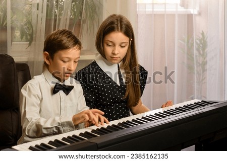 Dressed in formal attire, a young brother and sister concentrate on playing a piano duet in their living room Royalty-Free Stock Photo #2385162135