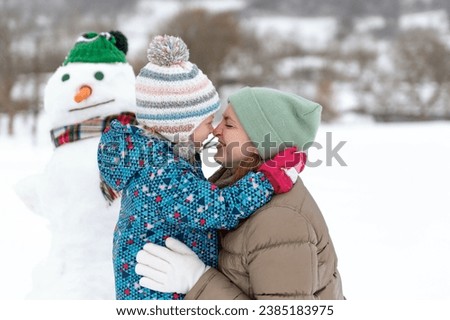 Child embracing mom against the snowman wearing hat and scarf. Portrait of mother and child in winter outside