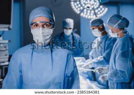 Portrait of female surgeon wearing scrubs and protective glasses in hospital operating theater. Serious surgeon doctor looking at camera in protective blue gown. Medical workers teamwork Royalty-Free Stock Photo #2385184381