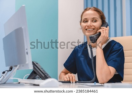 Smiling young woman nurse receptionist talking on phone while working in modern clinic. Making doctor`s appointment, consulting assisting helping customers patients clients in hospital Royalty-Free Stock Photo #2385184461