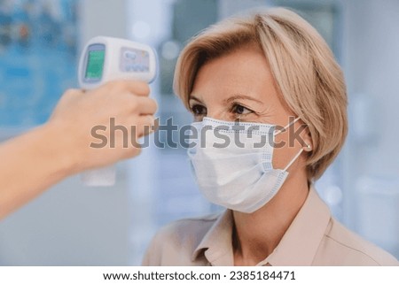 Receptionist checking fever middle age woman by digital thermometer for fever scan. Healthcare concept. New normal, social distancing, pandemic, coronavirus Royalty-Free Stock Photo #2385184471
