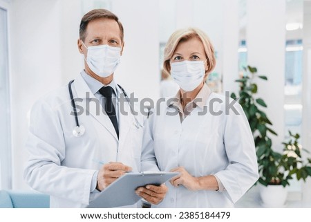 Adult female and male doctors standing together on clinic foyer while looking at camera wearing face masks for prevention epidemic. New normal. Social distance from Covid 19 Royalty-Free Stock Photo #2385184479
