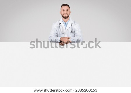 Young European male doctor standing confidently next to large blank placard board, ideal for medical ads on grey background, banner, copy space