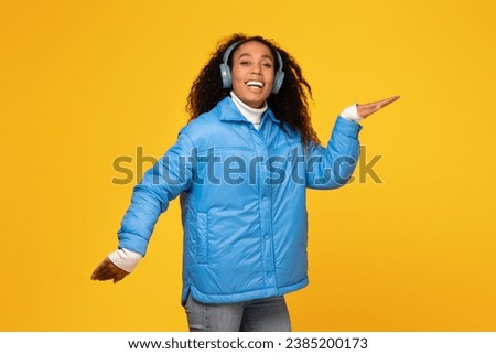 Joyful young black lady in winter jacket moves to music in wireless headphones, against yellow background, radiating cheerful holiday mood