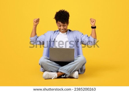 Exuberant young black man person sitting cross-legged, arms raised in victory, while looking at laptop computer on bright yellow background