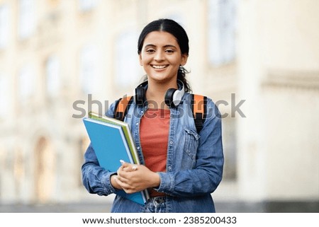 Positive young indian woman student wearing casual outfit with backpack and notepads exercise books posing outdoor at university campus, smiling at camera, copy space. Students lifestyle Royalty-Free Stock Photo #2385200433