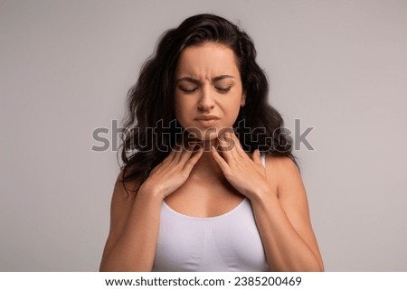 Sore throat, tonsilitis concept. Unhappy millennial brunette woman touching her neck, suffering from pain in throat, checking enlarged adenoids isolated on grey studio background, copy space Royalty-Free Stock Photo #2385200469