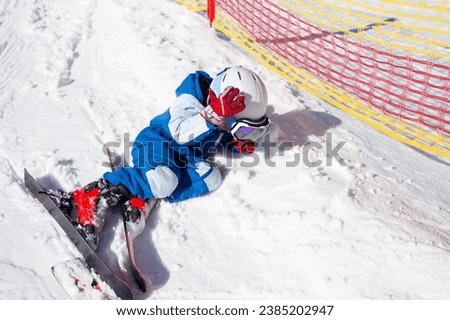 preschooler boy in a helmet, goggles, skis and winter overalls learns to ski, fell and lies on the snow. The tired child lay down on the snow to rest. Winter fun. winter holidays on a sunny cold day Royalty-Free Stock Photo #2385202947