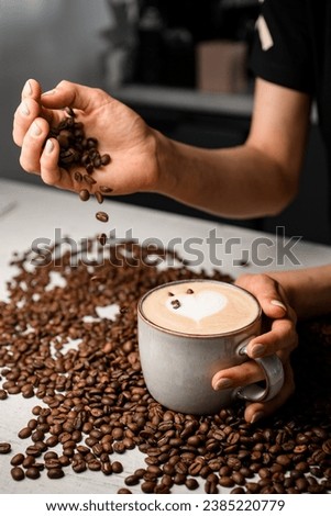 Selective focus of woman hands sprinkles roasted coffee beans to cup with freshly brewed cappuccino. Coffee beans on white kitchen table on background. Creative shot for coffee house