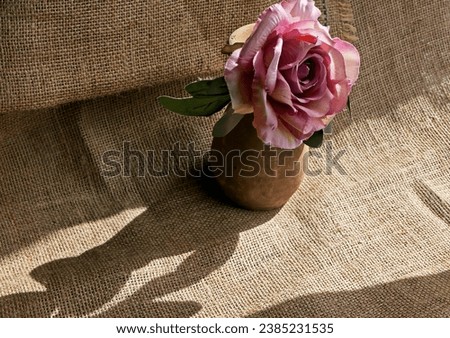 still life with artificial rose in a clay vase on burlap fabric background