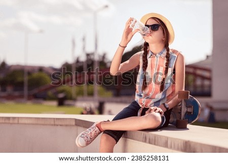 Teenager girl sitting on the bridge with skateboard and drinking water from a bottle