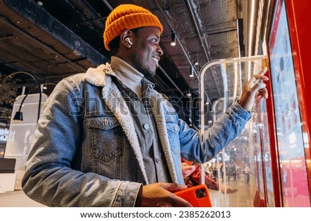 Smiling African-American man in warm denim jacket with wireless earphones uses self-service kiosk to order snack in cafe Royalty-Free Stock Photo #2385262013