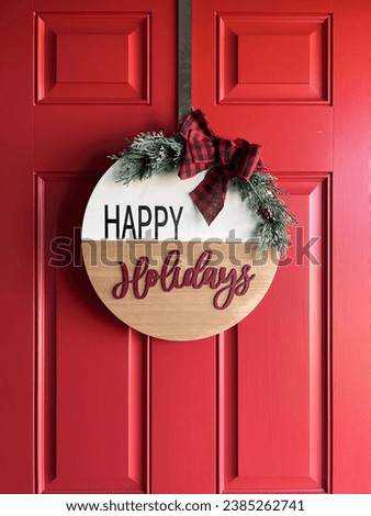 DIY craft style round wood welcome sign with woodgrain and white background and red cursive font reading "happy holidays" with a crafted farmhouse ribbon and green sprigs against a red door.