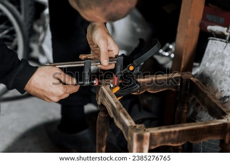 An adult male professional woodworker carpenter glues a chair, repairs, restores furniture indoors, workshop, clamping wooden products with a clamp. Photography, work concept, portrait.
