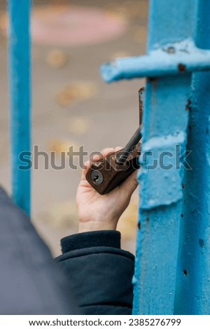 A child's hand holds an old padlock of a closed metal gate.