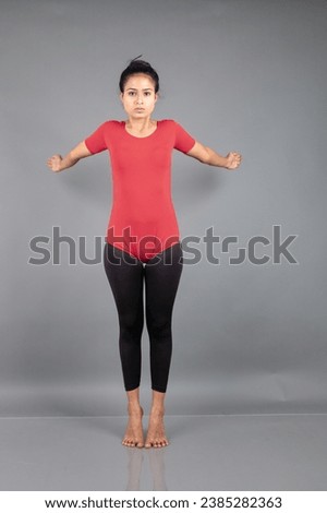 Healthy young woman in red t-shirt and black leggings doing yoga and stretching exercises isolated on grey background