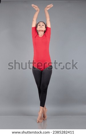 Healthy young woman in red t-shirt and black leggings doing yoga and stretching exercises isolated on grey background