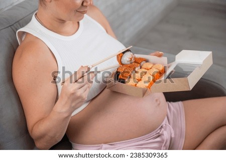 A pregnant woman sits on the sofa and eats rolls from a box. Food delivery.  Royalty-Free Stock Photo #2385309365