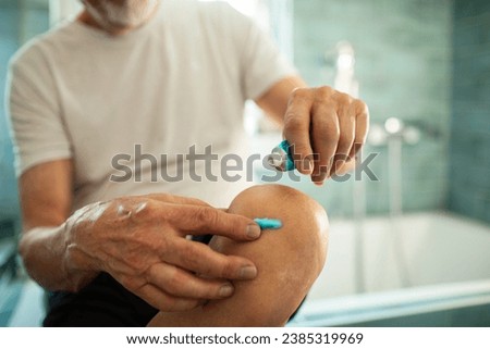 Senior Man Applying Ointment on Knee in Bathroom Royalty-Free Stock Photo #2385319969