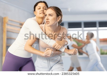Focused young woman practicing basic protection skills in mock bout with female rival during self-defense course in gym, trying to break free of rear grab, striking solar plexus with elbow Royalty-Free Stock Photo #2385325605