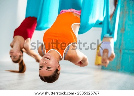 Smiling girl doing dove pose during aerial yoga group training in fitness room, lying in blue suspended hammock, arching and folding outstretched arms into lock behind her back..