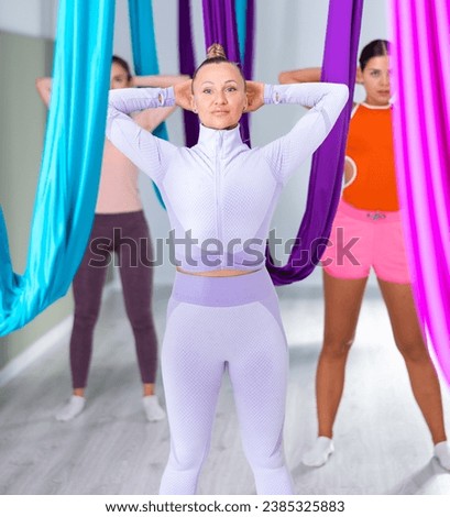 Active sporty adult woman doing articular gymnastics exercises before aerial yoga group training in fitness room among multi-colored silk hammocks