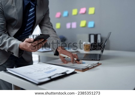Asian businessman using mobile phone checking financial market app, calculating, planning, analyzing data in documents. Chat online on a laptop computer in the company office.