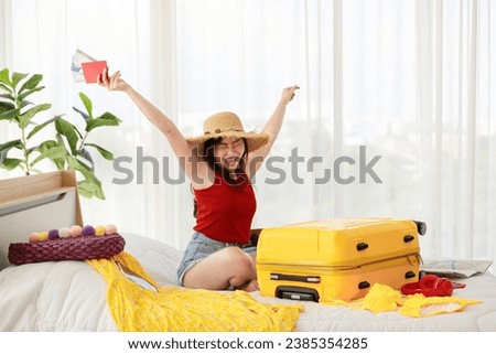 Asian young happy female traveler sitting on bed holding passport with air tickets with happy and excited actions while packing personal traveling stuff to yellow trolley baggage on bed.