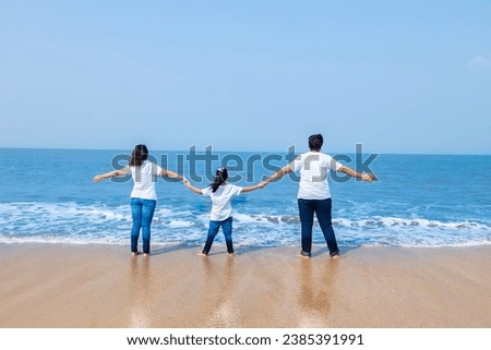 Happy Back view of young indian family wearing white t-shirt and blue jeans having fun at the beach standing with open arms. Summer vacation and travel concept