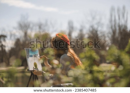 Close up of ginger girl with red hat painting a landscape while sitting at the park near the lake