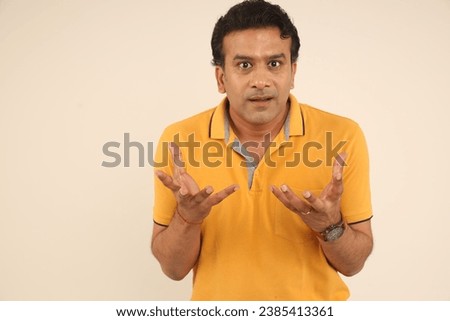 Young indian man wearing t-shirt standing over isolated studio background funny facial expressions, shocking puffing cheeks with funny face. Mouth inflated with air, crazy expression.
