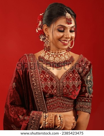 The image portrays a beautiful Indian woman dressed in a traditional red and green lehenga.
The woman is posing with a bright smile on her face.he background of the picture is a deep red color.