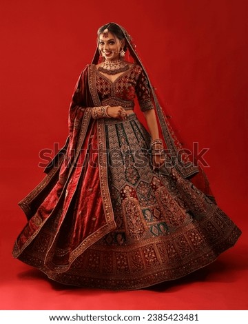 The image portrays a beautiful Indian woman dressed in a traditional red and green lehenga.
The woman is posing with a bright smile on her face.he background of the picture is a deep red color. Royalty-Free Stock Photo #2385423481