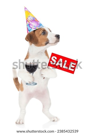 Jack russell terrier puppy  wearing a party cap holds glass of red wine and shows signboard with labeled "sale". Isolated on white background