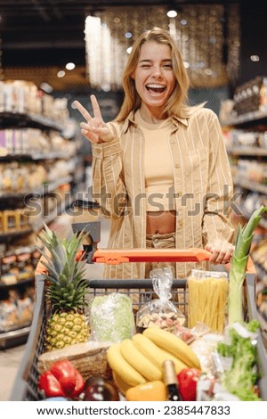 Young cool customer woman wear casual clothes show v-sign wink shopping at supermaket store grocery shop buying with trolley cart choose products inside hypermarket. Purchasing food gastronomy concept