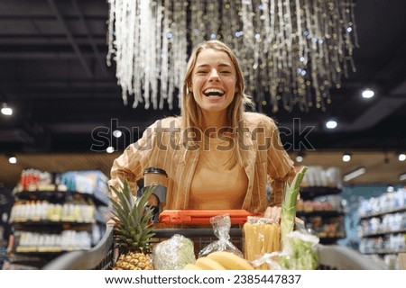 Young smiling customer woman wear casual clothes look camera shopping at supermaket store grocery shop buying with trolley cart choose products inside hypermarket. Purchasing food gastronomy concept