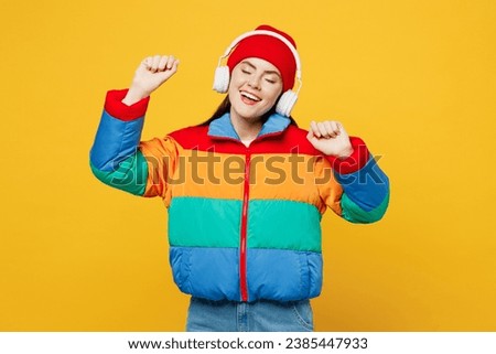 Young smiling happy woman she wears padded windbreaker jacket red hat casual clothes listen to music in headphones raise up hands dance isolated on plain yellow background studio. Lifestyle concept