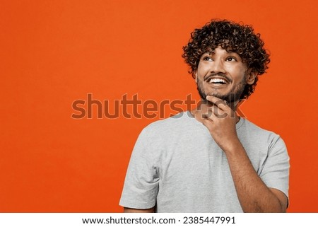 Young smiling happy Indian man he wearing t-shirt casual clothes put hand prop up on chin, lost in thought and conjectures isolated on orange red color background studio portrait. Lifestyle concept