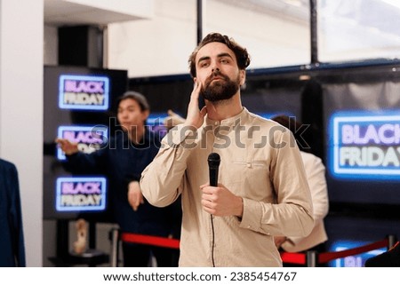 TV reporter live broadcasting talking about Black Friday lines in local shopping mall, holding microphone and looking at camera. Young man correspondent covering news about sales season in retail Royalty-Free Stock Photo #2385454767