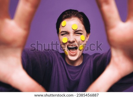 Quirky young woman taking a selfie with smiley stickers on her face. Playful woman smiling cheerfully while taking a picture of herself. Happy young woman having fun against a purple background.