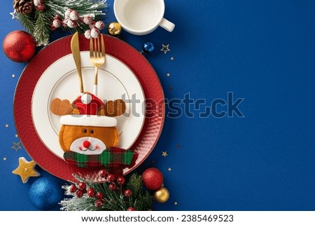 Cheerful New Year's kids' table decor. Top-view photo of plates, playful pocket utensil holder, mug, balls, candle, sparkle confetti, frosty fir twigs, and mistletoe berries on blue backdrop, ad space