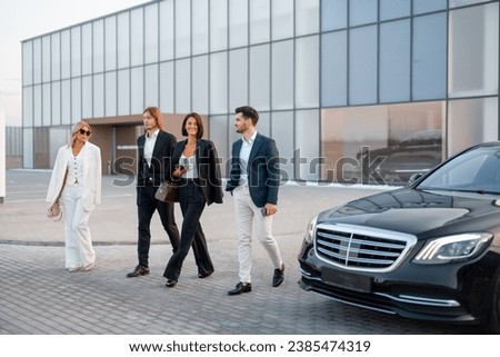 Group of business people talking on the go from modern building of airport or some office to luxury taxi on parking lot on sunset. Business partners are going to leave by luxury car Royalty-Free Stock Photo #2385474319