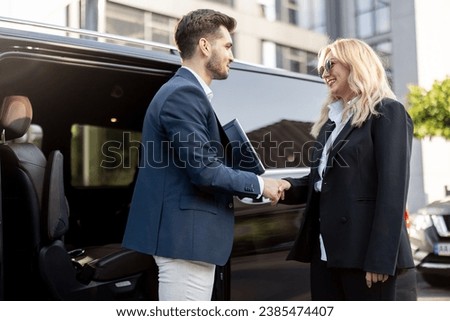 Female chauffeur greets the businessman while helping him to get out of the minivan taxi. Concept of personal driver, luxury taxi for business people Royalty-Free Stock Photo #2385474407