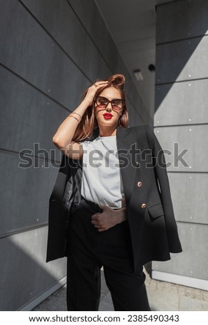 Stylish beautiful young woman with vintage sunglasses with red lips in fashion urban casual clothes with a blazer and T-shirt stands near a gray building with sunlight. Pretty business girl Royalty-Free Stock Photo #2385490343