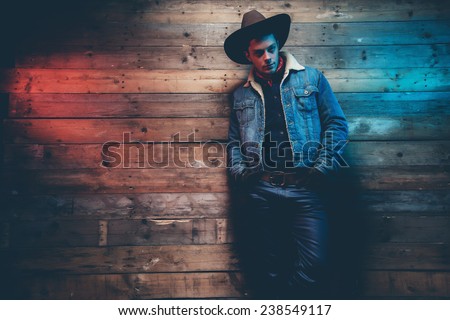 Winter cowboy jeans fashion man. Wearing brown hat, jeans jacket and trousers. Leaning against old wooden wall. Royalty-Free Stock Photo #238549117